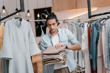 young business owner holding pile of pants while working in boutique before opening