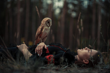Girl lies with owl on grass in forest.