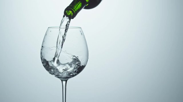 Closeup of mineral water being poured from green glass bottle into wineglass against white background