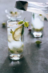 A glass of lemonade with ice, a pouring syrup, opposite the light, from lemon, gooseberries, rosemary, on a concrete background