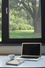 Fototapeta premium Laptop computer, notepad with pencil and cup of coffee on white desk in front of window at home office