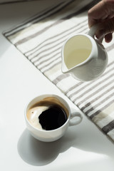 Person pouring milk from jug into cup of black coffee on white table with tablecloth