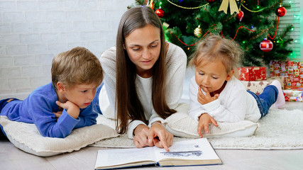 Happy Mother with two childes lying on the floor and reading book at xmas eve.