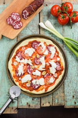 Raw rustic pizza with smoked salami, tomatoes, green olives and cheese served with pizza knife and ingredients: leek, cherry tomatoes and salami