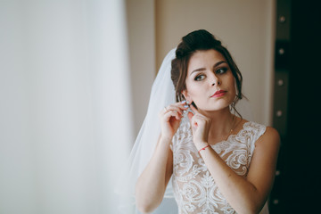 Beautiful bride puts on earrings at the window