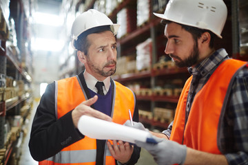 Portrait of warehouse worker talking to supervising manager while doing inventory control in...