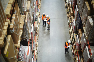Above view of warehouse workers group in aisle between rows of tall shelves full of packed boxes...