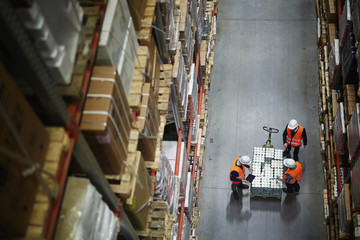 Above view of people working in large warehouse, counting goods on moving cart between shelves with...