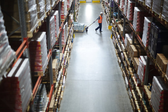 Side view of warehouse worker pulling moving cart between tall shelves with packed boxes and goods