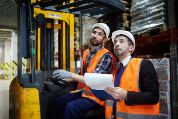 Portrait of warehouse manager and loader riding forklift truck inventorying stock