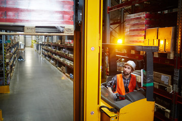 High angle portrait of warehouse worker using reach fork truck to load pallet with boxes on tall rack