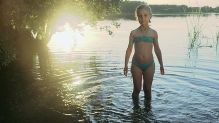 Cute little girl is splashes and catches soap bubbles standing in the water of a picturesque forest lake in the light of a glares on the water. Slow motion. - Powered by Adobe