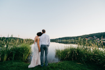 Happy bride and groom after wedding ceremony standing at shore of lake in evening at sunset on nature. Young married couple taking hands and lookig at horizon. Romantic view. Marriage celebration.