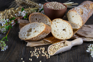 Bakery concept. Buckwheat bread, French baguette and stalks of whea, oats, buckwheat on dark wooden table.