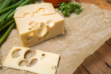 mouthwatering delicious piece of maasdam cheese and a slice on a wooden table surrounded by greenery and tomatoes