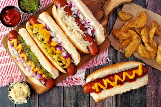 Hot dogs with assorted toppings and potato wedges, above view on a rustic wood background