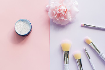 Five makeup brushes with lettering on the handle and mineral powder in a blue jar, bobby pin in the form of a pink flower on pink and purple background. Have copy space.