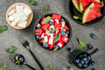 Summer fruit berry salad: watermelon, blueberries, mint, feta cheese and balsamic sauce. On a black bowl, black stone table. With ingredients, fork, knife. Copy space top view