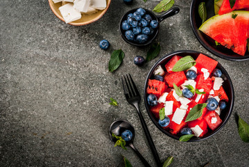 Fototapeta na wymiar Summer fruit berry salad: watermelon, blueberries, mint, feta cheese and balsamic sauce. On a black bowl, black stone table. With ingredients, fork, knife. Copy space top view