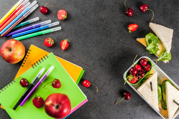 Back to school. A hearty healthy school lunch in a box: sandwiches with vegetables and cheese, berries and fruits (apples) with notebooks, colored pens on a black table. Copy space top view