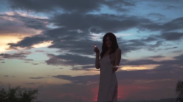 beautiful sexy girl in vecherom dress dancing on the beach at sunset. a young woman holds the sparklers and have fun at the party. slow motion
