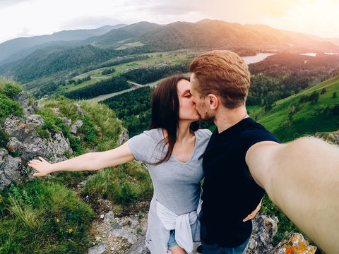 Guy and girl tourists are happy, hug and do selfie on action camera on background of mountains, forests. Concept kiss and travel. Russia, Altai Mountains.