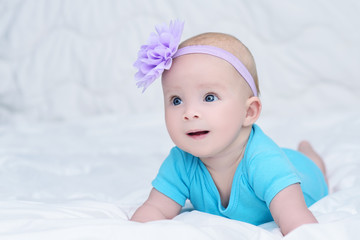 Cute baby girl in blue shirt, with bow flower on head, lying on stomach on soft blanket and looking with surprise, indoors