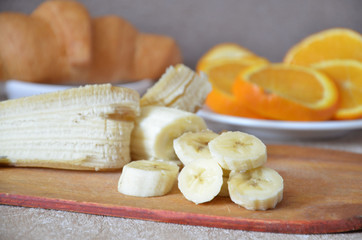 Pieces of banana cutting board chopped with a knife. Sliced orange and croissant on background