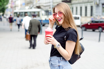 Outdoor portrait of young attractive woman with long blonde straight hair, pink glasses,  and berry's smoothie cocktail, street urban background. Healthy food detox concept. Windy hair flip