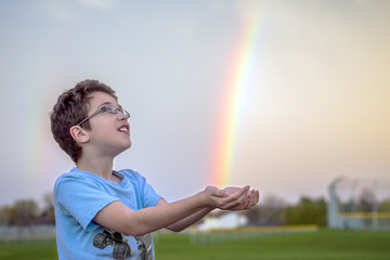 Young boy looking up in awe at bright rainbow after spring rain, trying to catch the colors in his hands