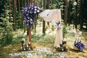 Wedding wooden arch for marriage ceremony with flowers, curtain and other decoration elements standing on ground in forest. Pine trees in sunny summer day. Celebration mood. Candles in glass, feathers