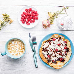 Homemade pancakes or Russian pancakes with chocolate sauce, whipped cream and raspberries on a plate on a white wooden background.