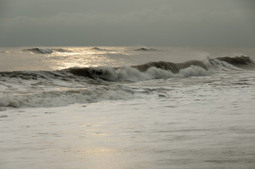 A long powerful brown wave run to the coastline, weather is stormy, sky is low and grey, picture is dramatic.