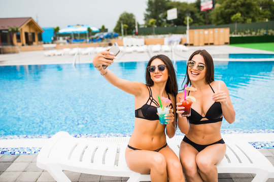 Two attractive brunette women wearing bikini posing near the swimming pool, making selfie photo. Summer time