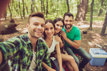 Close up of four happy friends tourists in the spring nice wood, embracing, posing for a selfie shot, that handsome blond guy is taking, sitting on a log