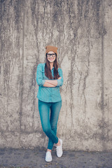 Obraz na płótnie Canvas Full length of excited girl, standing on the concrete wall`s background outdoors, smiling, with crossed legs and hands, in casual jeans outfit and brown hat, glasses