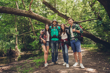 Close up of four cheerful  friends in the summer nice wood. They are hikers, walking and picking place for camping, embracing, posing for photo with equipment