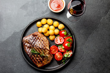 Grilled beef steak served on cast iron plate with tomato salad, potatoes balls and red wine. Barbecue, bbq meat beef tenderloin. Top view, slate background