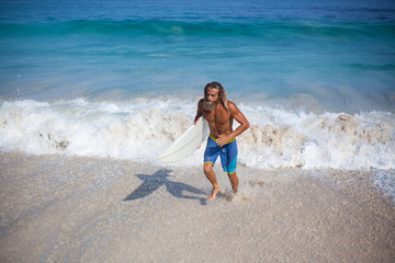 Horizontal portrait of a handsome bearded curly wet man with naked torso with white surfboard runing out of the ocean