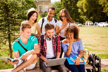 It`s so fun and easier together! Six happy multi ethnic students are learning outdoors in a sunny park, with books, note pads and devices, dressed comfortable, laughing