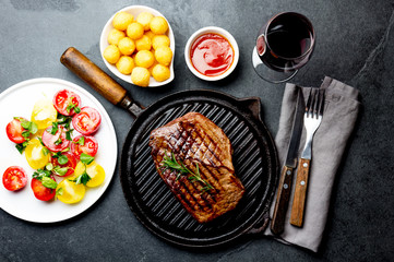 Grilled beef steak on grill pan served with tomato salad, potatoes balls and wine. Barbecue, bbq meat beef tenderloin. Top view, slate background