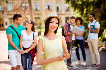 Fototapeta premium Young attractive asian student is holding tablet. She is wearing casual smart clothes, hat, behind her are her classmates, in a sunny park near the building of campus