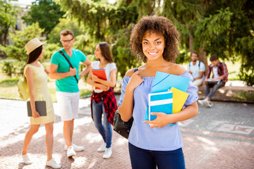 Fototapeta na wymiar Portrait of an attractive young student, standing with rucsack and note pads, with beaming smile, behind are her friends, talking outside in a sunny park near college