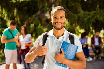 Young cheerful successful african nerdy student is showing thumb up, standing with books, his classmates on the background, park near campus, sunny day