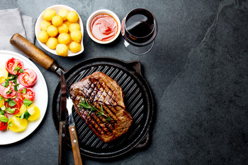 Grilled beef steak on grill pan served with tomato salad, potatoes balls and wine. Barbecue, bbq meat beef tenderloin. Top view, slate background