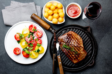 Grilled beef steak on grill pan served with tomato salad, potatoes balls and wine. Barbecue, bbq meat beef tenderloin. Top view, slate background