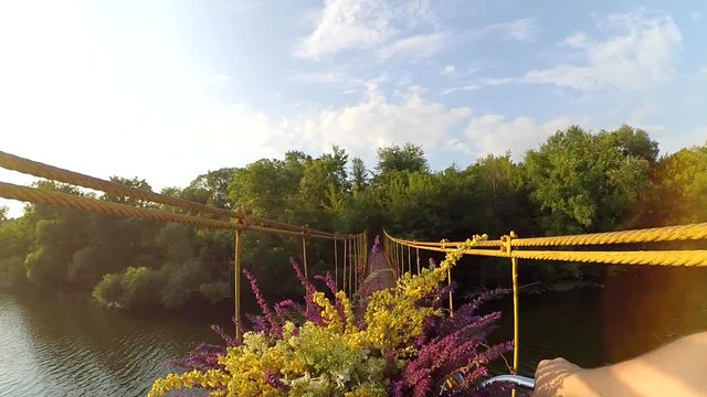 Slown motion. A girl on a bicycle with a bouquet of multicolored wildflowers in the corms before the wheel passes over a suspension bridge. Action camera go pro.