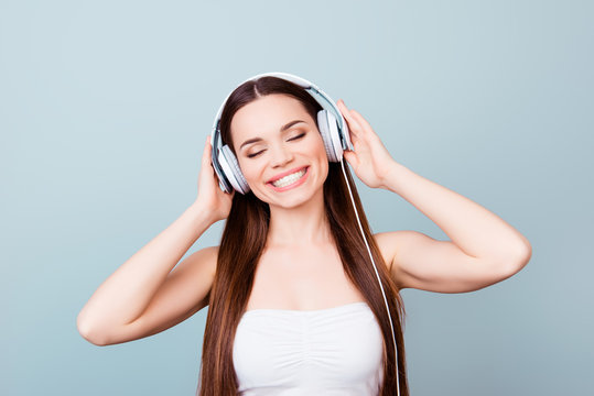 Charming young girl with toothy smile in modern headphones. She is dreamy and happy, listening to her favourite song, holding the earphones, with closed eyes on pure background