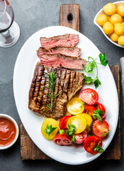 Sliced medium rare grilled beef steak served on white plate with tomato salad and potatoes balls. Barbecue, bbq meat beef tenderloin. Top view, slate background, copy space