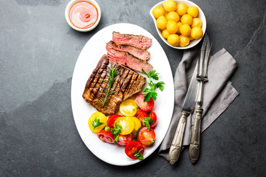 Sliced medium rare grilled beef steak served on white plate with tomato salad and potatoes balls. Barbecue, bbq meat beef tenderloin. Top view, slate background, copy space